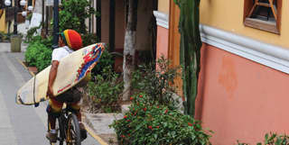 A man in a pan-African green, yellow and red hat, cycles with a surfboard under one arm down a bright street in Lima.