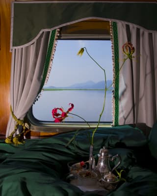 A tray with a silver tea set rests on a green duvet adorned with a flame lily that stands out against the misty window view.