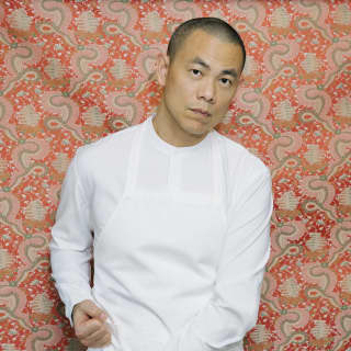 Chef Andre Chiang stands against a red and green-patterned wall, tightening his white apron straps while looking to camera.