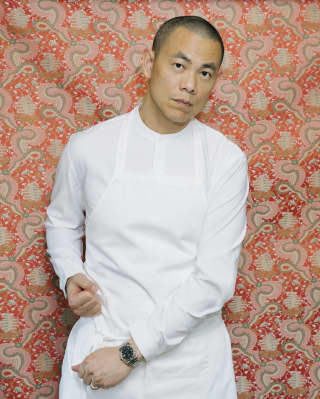 Chef Andre Chiang stands against a red and green-patterned wall, tightening his white apron straps while looking to camera.