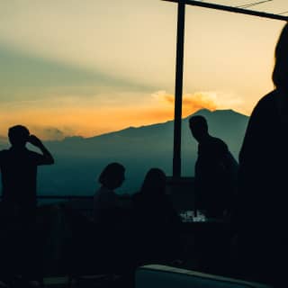 Silhouetted guests watch the horizon where Mount Eta rises dramatically, plumes of smoke tinged apricot by the sunset above.