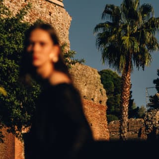 An out-of-focus woman in the foreground stands in front of stone ruins and a palm tree, crisp and clear in bright sunlight.