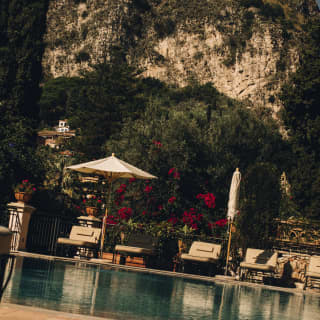 Ground-level view over the pool, where loungers recline beneath two parasols, one folded, with a dramatic mountain backdrop.