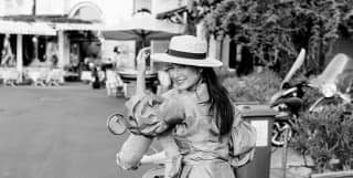 A young women holds onto her straw boater and smiles back at the camera as she rides pillion on a Piaggio scooter