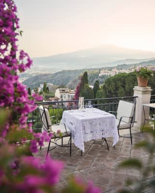 Bougainvillea tumbles down, framing the view of a lace topped terrace dining table for two, with Mount Etna rising behind