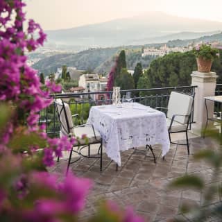 Bougainvillea tumbles down, framing the view of a lace topped terrace dining table for two, with Mount Etna rising behind