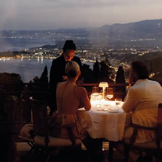 A waiter stands by a couple at a lamp-lit dining table at Otto Geleng with sweeping dusk views of the light-studded coast.