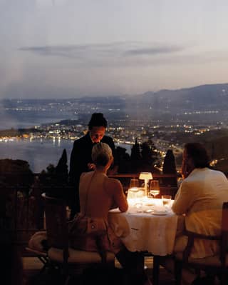 A waiter stands by a couple at a lamp-lit dining table at Otto Geleng with sweeping dusk views of the light-studded coast.