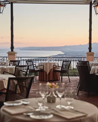 Formal linen-coated table in the foreground of a restaurant terrace with views of Taormina at dusk beyond