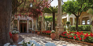 Bougainvillea drapes from an iron pergola shading a large doorway. The entrance is lined by red geraniums and white petunias