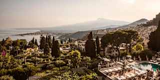 View over gardens with shrubs and trees and a lounger-lined pool, down to Taormina's coastline and Etna's distant silhouette.