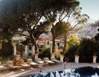 High angle view over the pool edge where water captures the reflection of pines, under which two woman chat on sun loungers.