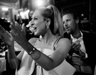 A woman with long blond hair in a ponytail, and a suited male companion behind, clap in celebration at a special occasion.