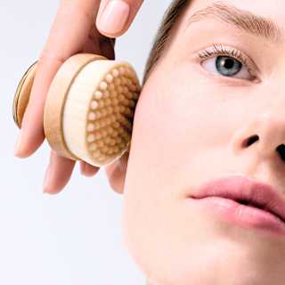 A hand holds a round lymphatic bristle brush against the cheek of a woman with natural-look make-up, in a Dior Spa image.