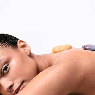 A woman with slicked-back dark hair lies on her front, looking to camera, with lemon and grey-toned stones on her back.