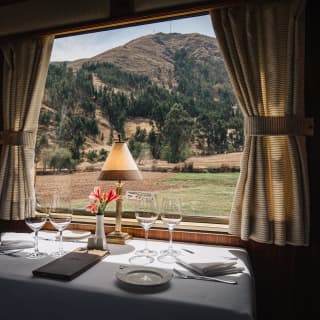 An elegant table laid for dinner looks through a train window bordered by curtains to hilly landscape in greens and browns.