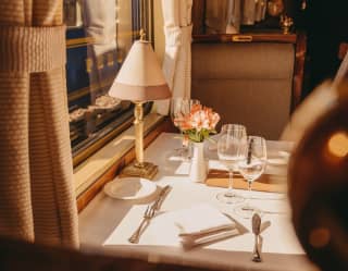 A dining table dressed with a white tablecloth, cutlery and glasses, adorned with a pink bloom in a vase, waits for guests.