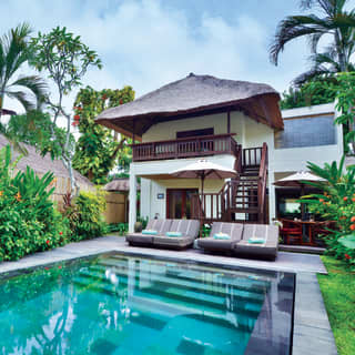 Looking across a crystal pool bordered by tropical plants to four sun loungers at the rear of a two-storey Garden View Suite.