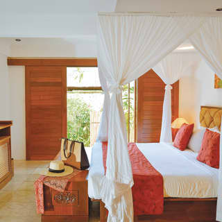 In a villa bedroom, a hat and bag rest on a box at the end of a bed with drapes tied back to reveal red cushions and a
throw.