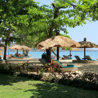 Blue-cushioned loungers recline beneath thatched parasols in dappled light on Jimbaran Beach, seen from the resort gardens.