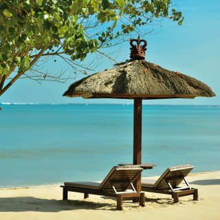 Either side of a straw parasol, two sun-chairs on the sand by the sea look out over the blue, alone but for a Cordia tree.