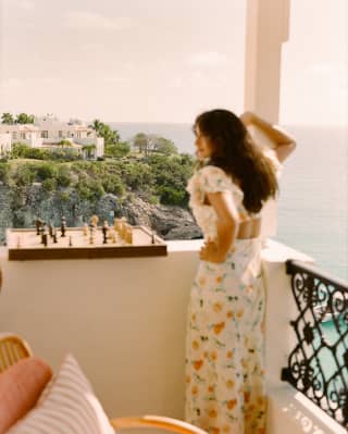 A woman with long dark hair and a floral dress leans with one arm on a balcony pillar, where a chess board rests on the wall.
