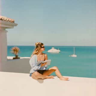 A woman with long, blond hair reads a book in a swimming costume and long baggy shirt, on a terrace wall overlooking the sea.