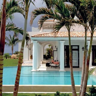 Palm trees offer shade and privacy to an infinity pool that wraps around the corner of a villa veranda supported by pillars