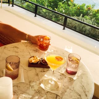 At Baie Longue Bar, a guest's hand reaches for a tumbler at a table with aperitifs and cocktails, with treetop and sea views.