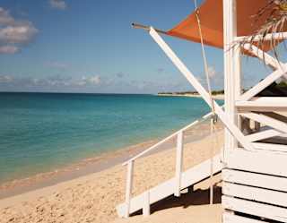 Side-view of a white wooden cabana with an orange shade sail and ramp onto the soft sand at the edge of the turquoise sea.