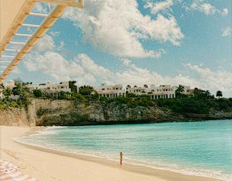 A figure stands at the edge of the sapphire sea, seen from beneath a canopy in a sun-drenched view of the beach and cliffs.