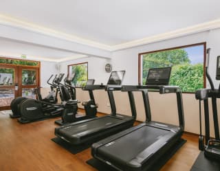 Six exercise machines - rowing, cycle, treadmill and crossfit - line a wall with two garden windows in the wood-floored gym.