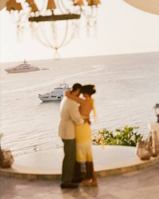 A man in a pale jacket embraces a woman in a yellow dress in the lobby overlooking two boats anchored in the evening ocean.