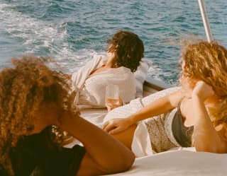 Three friends reclining on the cream, padded deck at the back of a boat watch the wake bubbling on the surface of the sea.