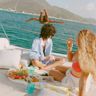 Two women relaxing and snacking on the deck turn to watch their friend diving from the boat, her leap captured in mid-air.
