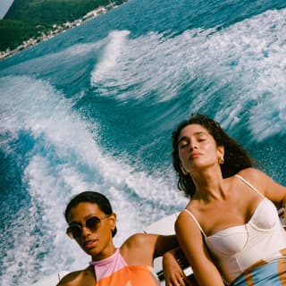 Two dark-haired women, one with hair back, the other down, bask in sun and spray as their boat speeds through churned waters.
