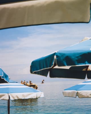 Action shot as a boy leaps into the sea from a rock where his friends sit in sunshine, seen through layers of blue parasols.