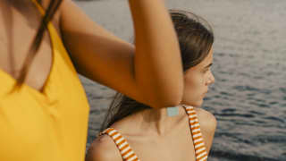 Two women in swimsuits stand on rocks at the water's edge. One clutches a straw hat as she gazes out over the Mediterranean
