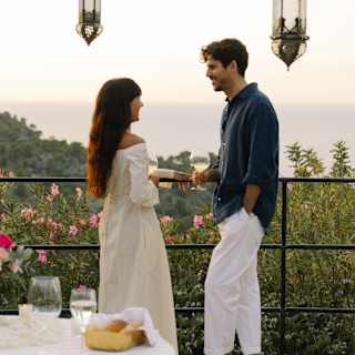 A woman in an ivory dress and a man in white trousers and black top drink and chat on the flower-filled sea-view terrace.