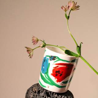 Angled image of a ceramic pot, painted with leaves and red and blue flowers in a composition with stones and a flower stem.