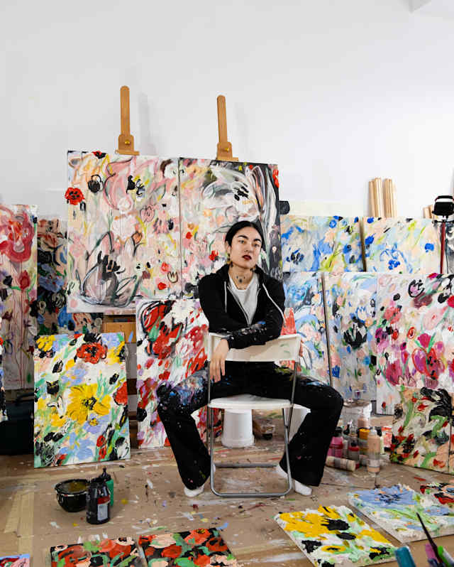 Artist Anna Deller-Yee, in black, sits astride a chair, arms on the backrest in a studio filled with vibrant floral artworks.