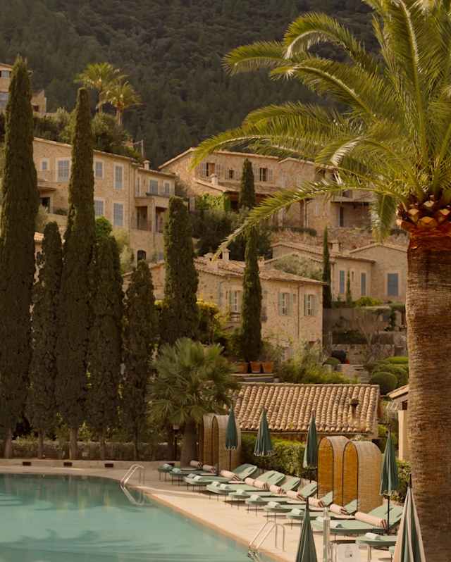The rustic hillside buildings of La Residencia rise behind a row of pencil pines, seen over a lounger-lined tropical pool.