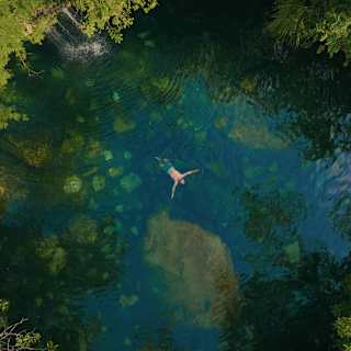 A man in trunks floats on his back in a beautiful, clear, cool-looking natural pool fed by a small waterfall, seen from above.