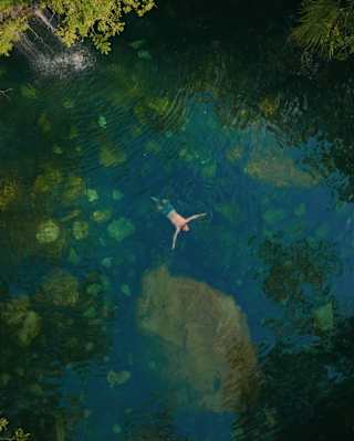 A man in trunks floats on his back in a beautiful, clear, cool-looking natural pool fed by a small waterfall, seen from above.