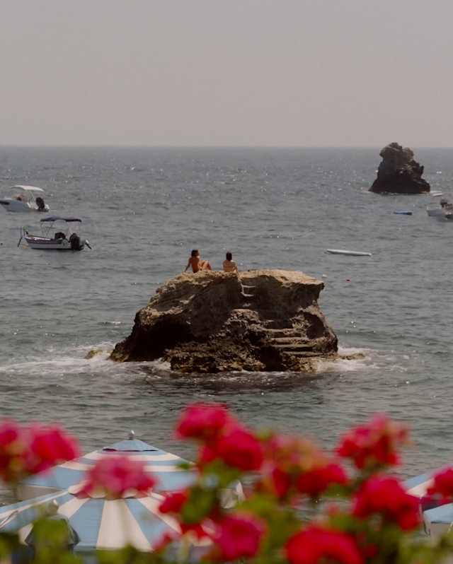 A couple sits atop a sea stack with carved rock steps, seen in a view over red geraniums, parasol tops and a sea of touring boats.