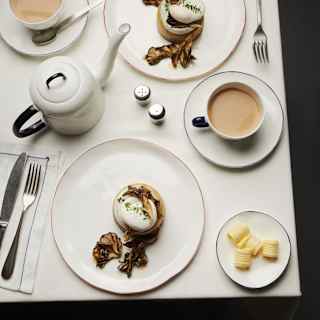 View down on two breakfasts of eggs on crumpets, with a dish of butter, a teapot and cups of tea, all served in white crockery.