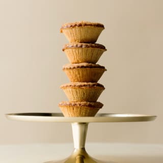 Close-up photograph of five baked pastry mince pies stacked in a tower on a silver cake stand, seen from the side.