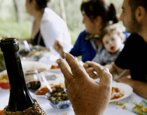 A bottle of wine in a raffia cooler rests on the edge of a dining table, where a family is enjoying a Mediterranean lunch.