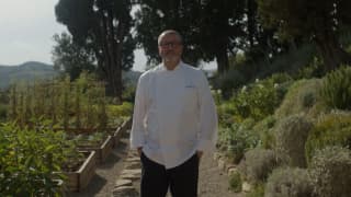 Chef Daniele Sera, with glasses, stands hands-in-pockets in an abundant kitchen garden between raised beds and herb borders.