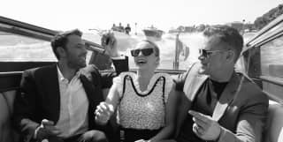 Black and white image of actors Ben Affleck, Jodie Comer and Matt Damon, dressed up and laughing together in a water taxi.